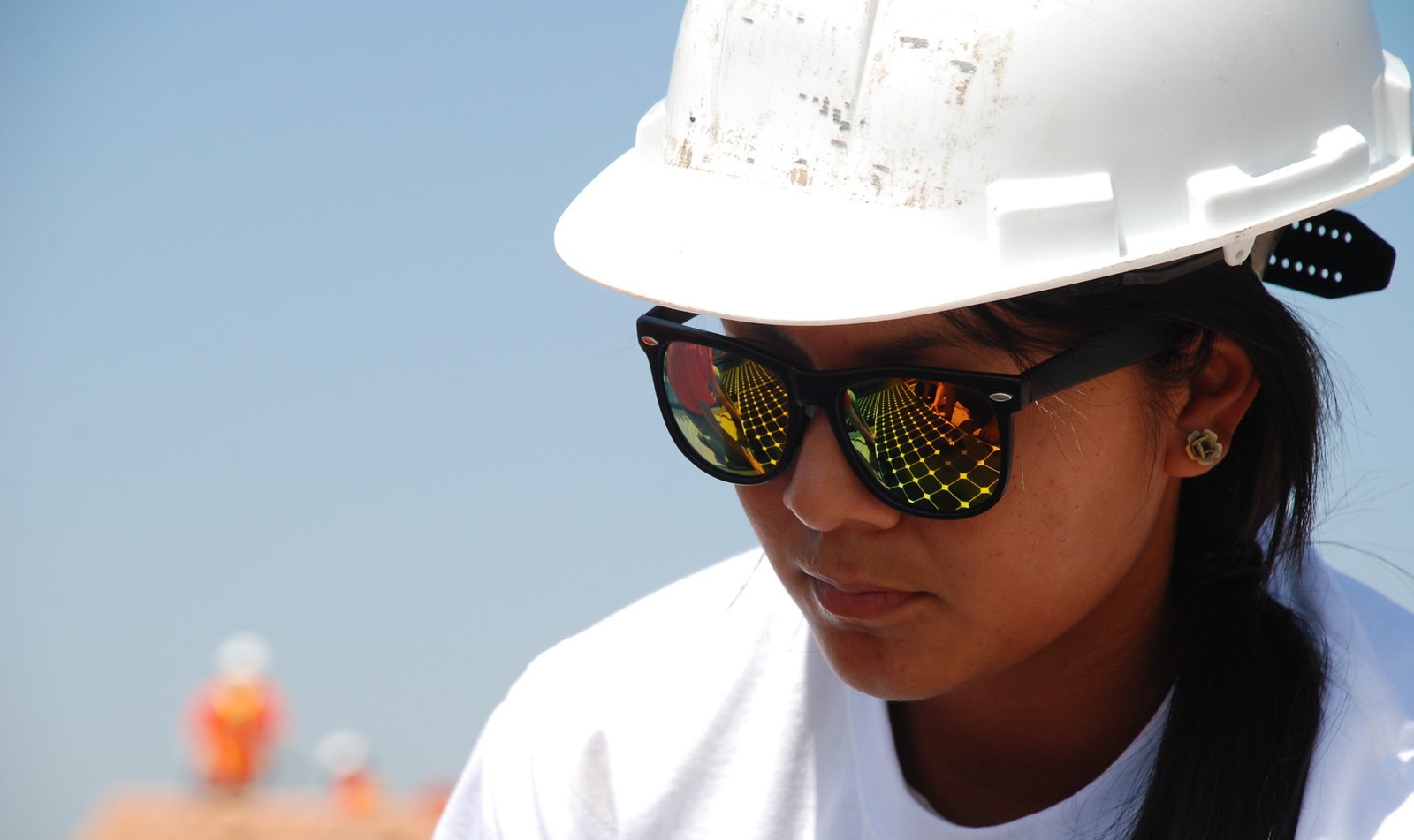 Close up of a woman wearing a white hard hat. Solar panels can be seen in the reflection of her sunglasses.