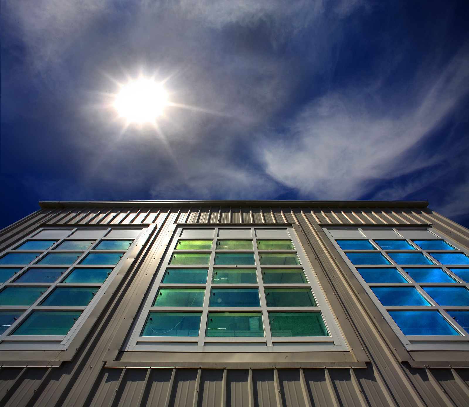 Low-angle shot of the Sun beating down on a tan, ridged building with three energy efficient windows. 