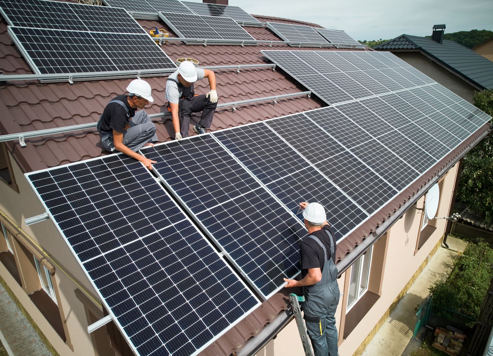 Three workers in grey overalls and white hard hats install a solar array onto a residential home's roof.