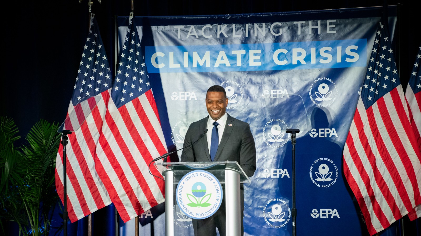 Administrator Regan smiling as he announces EPA's new power plant carbon rules at a podium. Beside him are American flags and behind him is a navy blue banner that reads 