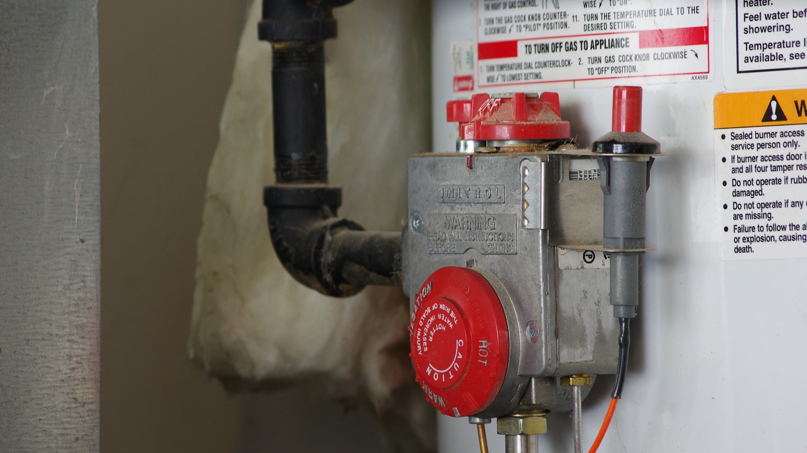 Close-up of a gas-fired water heater's red temperature controls and black supply pipe.
