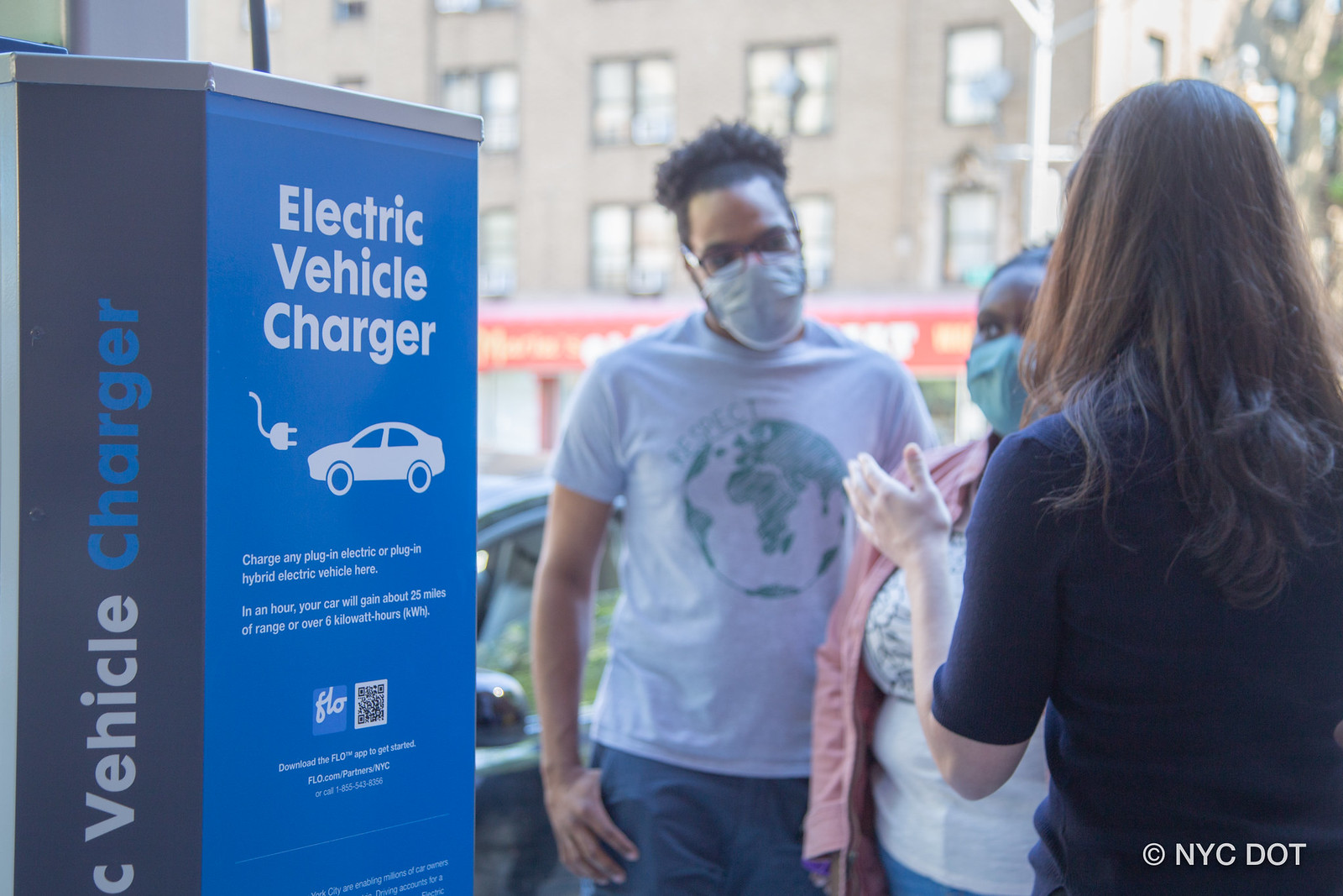 Community members listening intently to an outreach worker speaking. They are standing beside an electric vehicle charger.