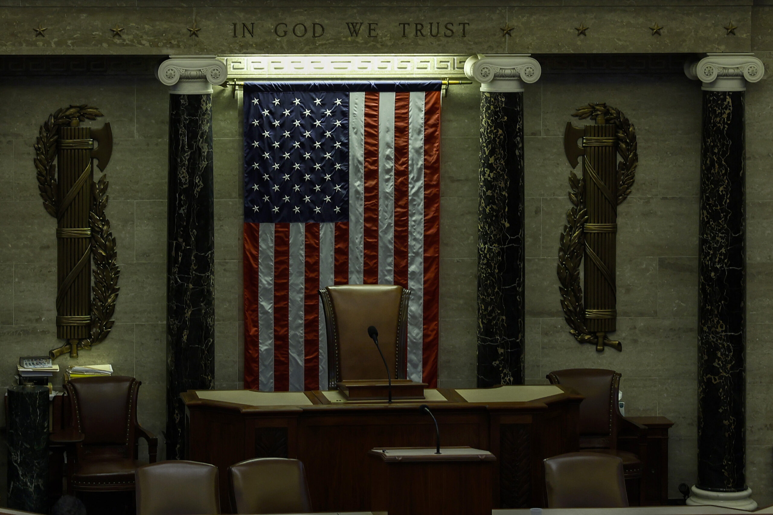 A picture of the front of an empty House of Representatives with the American flag hanging behind the podium and the words 