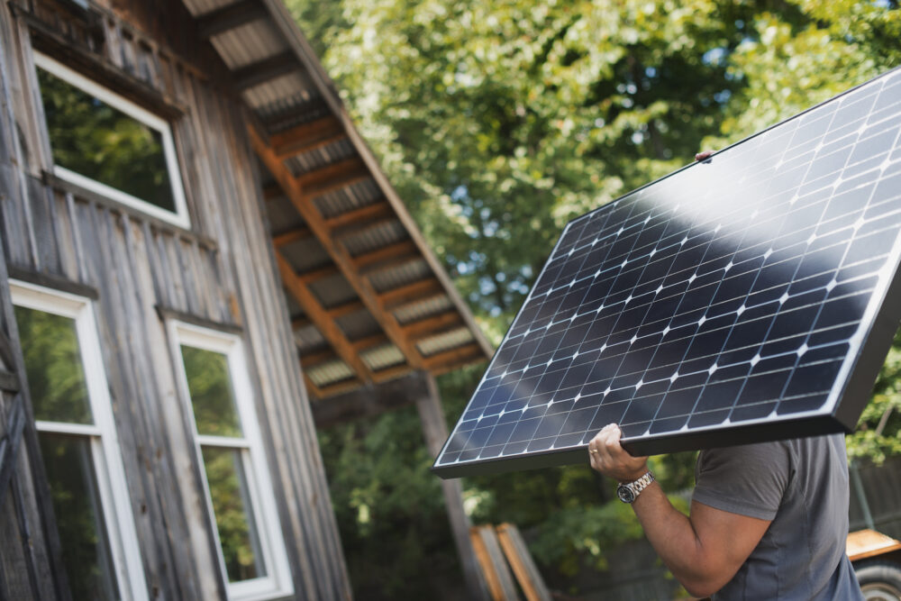 A person carries a solar panel on their shoulder near a wooden house surrounded by trees.