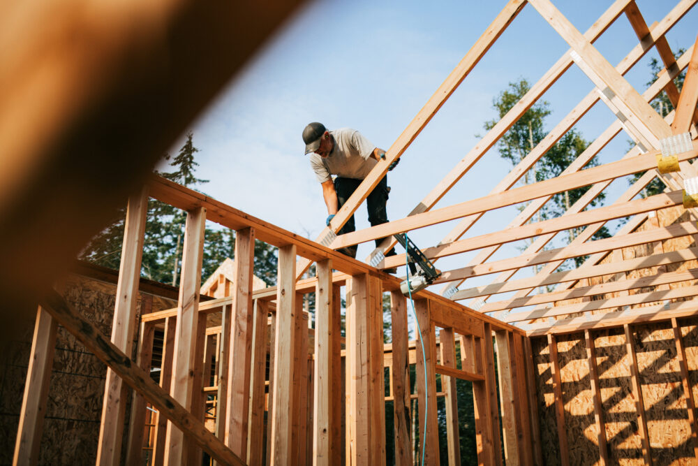 A construction worker balances on the wooden frame of a house under construction, positioning a roof truss beam, with trees visible in the background.