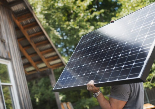 A person carries a solar panel on their shoulder near a wooden house surrounded by trees.