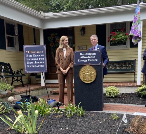 New Jersey Governor Mikie Sherrill speaks at a podium outside a home, alongside Assemblyman Bailey, at a press event about affordable energy, with signage reading '$1 Billion+ Per Year in Savings for New Jersey Ratepayers' and 'Building an Affordable Energy Future.'