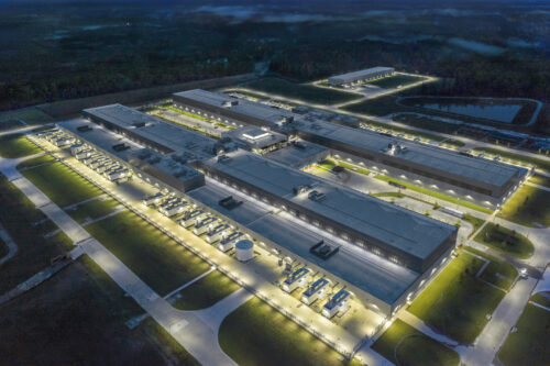 An aerial nighttime view of a large illuminated data center complex with rows of industrial cooling and backup power equipment, surrounded by forest.