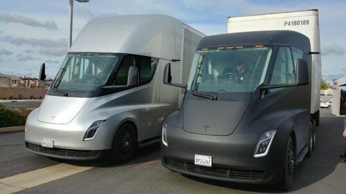 Two Tesla Semi prototypes at a Supercharging station in 2018. The silver truck (on the left) includes a large, removable aerodynamic cap on its roof, while the matte black truck does not.